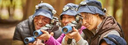 Laser-tag players aim blaster-style guns while smiling and focusing on a target; wearing bucket hats with head sensors and jackets; grouped closely in a sunlit forest setting.