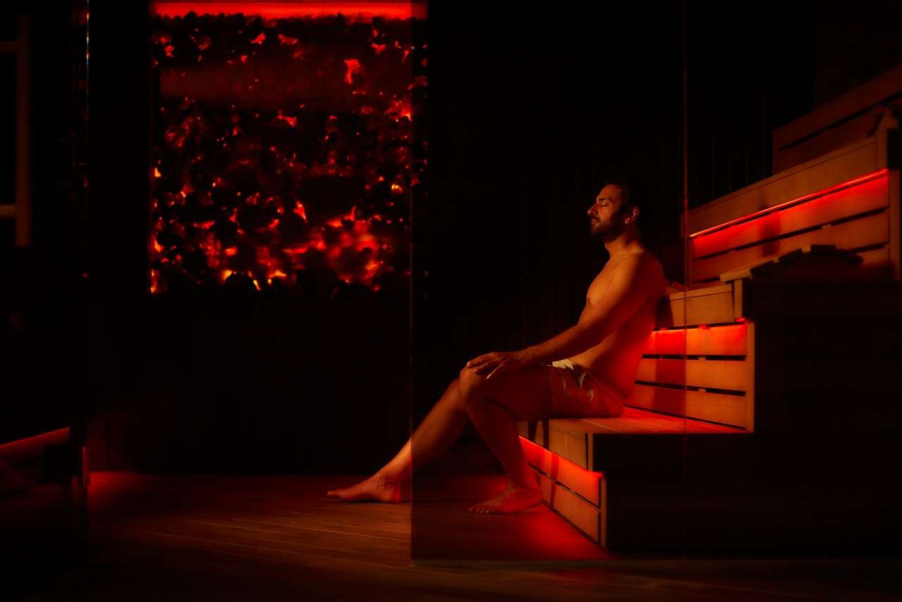 Person sits relaxing, eyes closed, on tiered wooden sauna bench; red ambient lighting glows from under seats and a backlit panel, surrounding a dim, enclosed spa room.