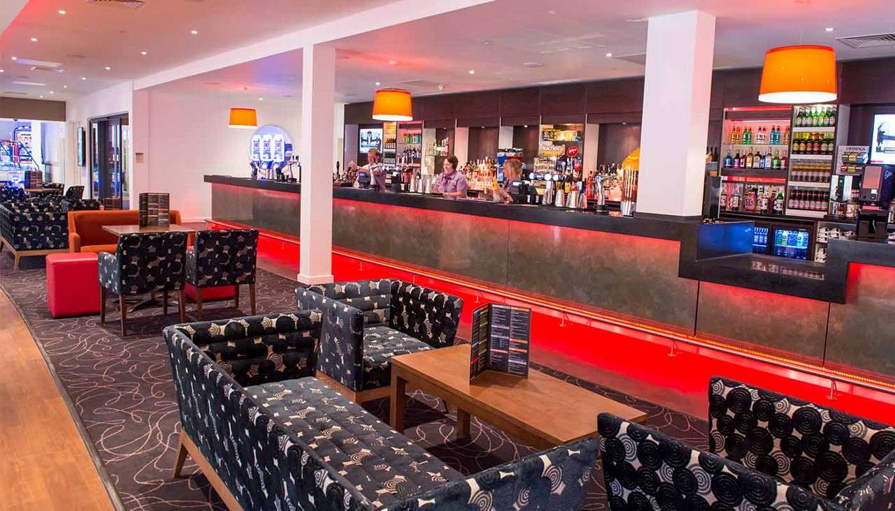 Bartenders serve drinks at a long illuminated bar, while patrons sit or stand nearby. Patterned armchairs and low tables fill a modern lounge with orange pendant lamps and stocked shelves.