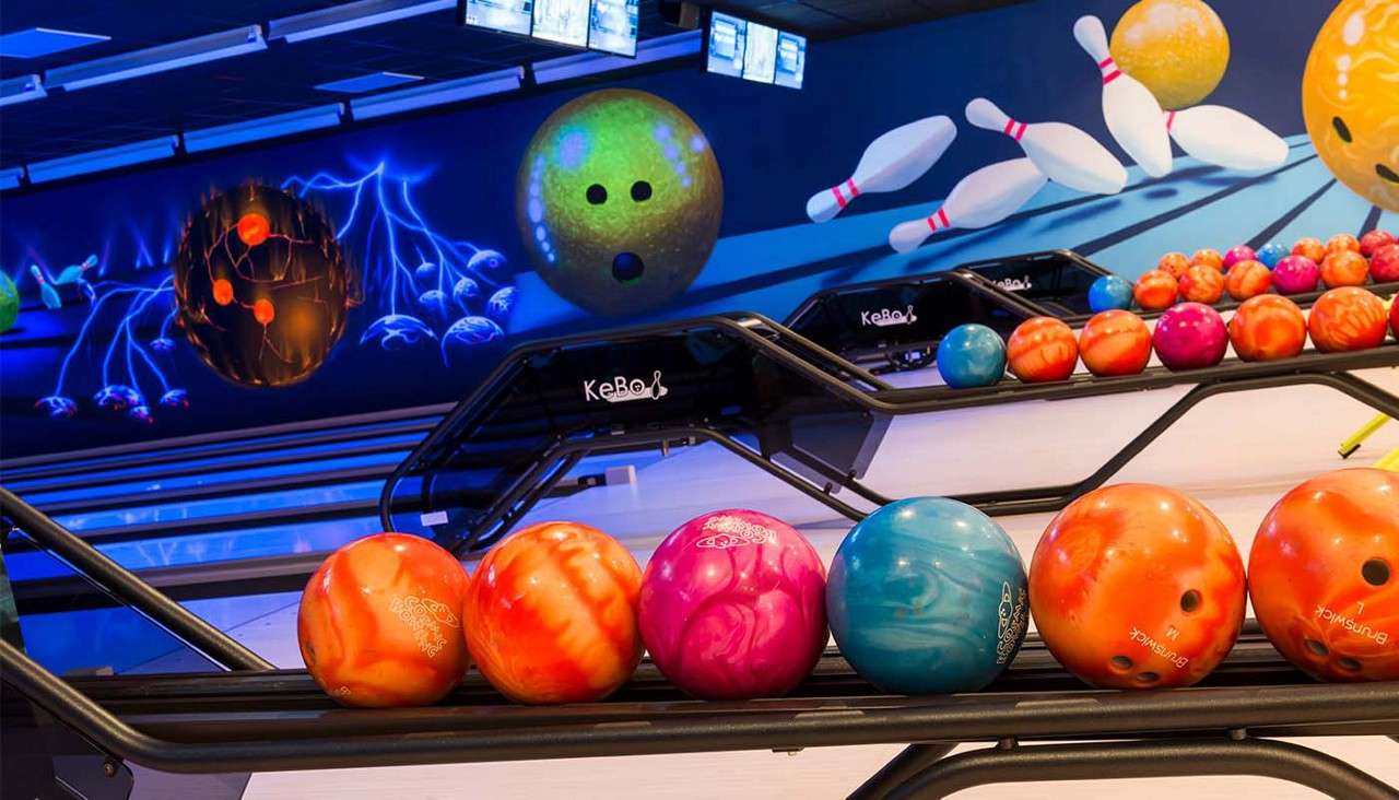 Colorful bowling balls line metal return racks, awaiting play; behind them, glossy lanes, overhead screens, and a neon mural of giant balls and flying pins. Text visible: KeBo; BRUNSWICK.