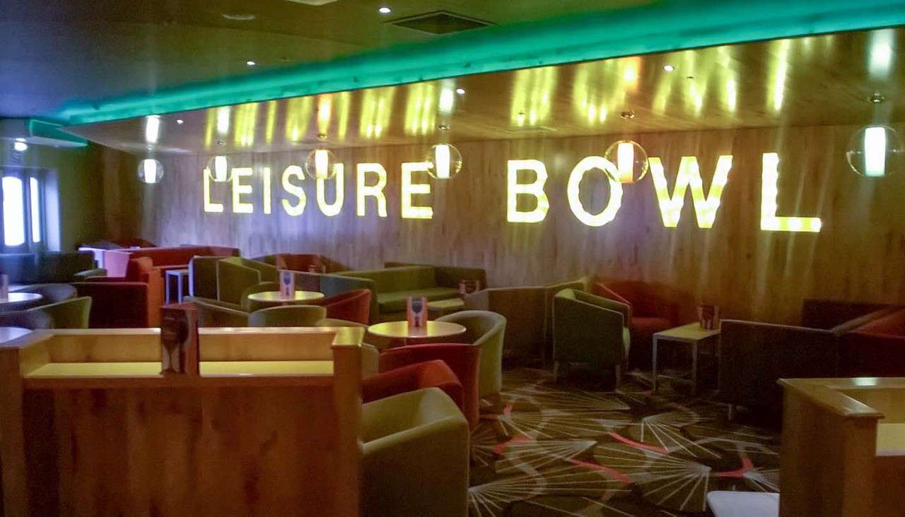 Illuminated marquee letters spell LEISURE BOWL, glowing on a wood‑paneled wall; upholstered chairs and small round tables sit arranged in clusters within a dim, modern lounge.