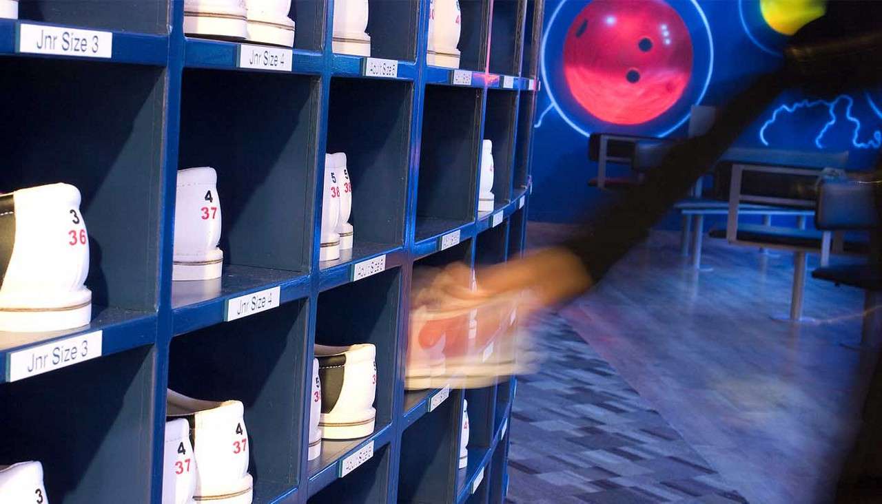 Bowling shoes rest in blue cubbies as a blurred hand grabs a pair; neon bowling-alley seating and murals behind. Visible text: "Jnr Size 3", "Jnr Size 4", "3 36", "4 37", "5 38".