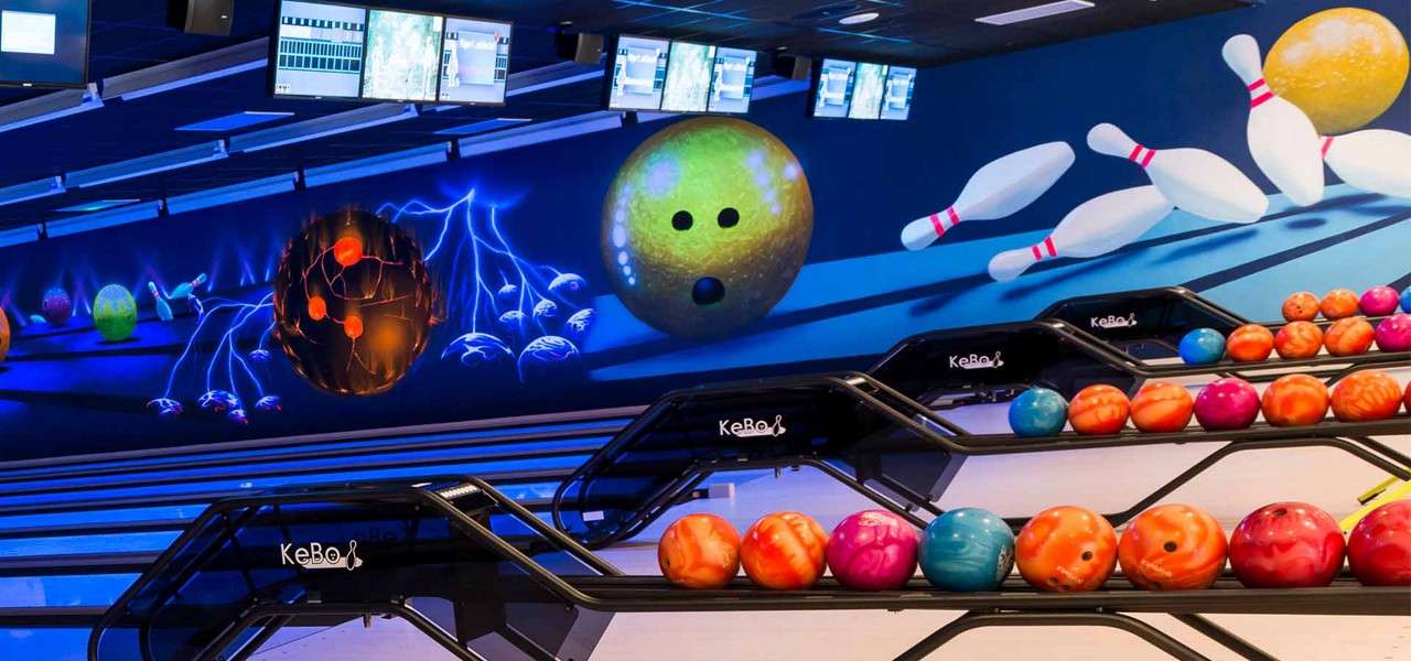 Bowling balls rest on racks, awaiting players, in a neon bowling alley with a mural of oversized balls and pins, overhead scoring screens, and rack branding text: KeBo.