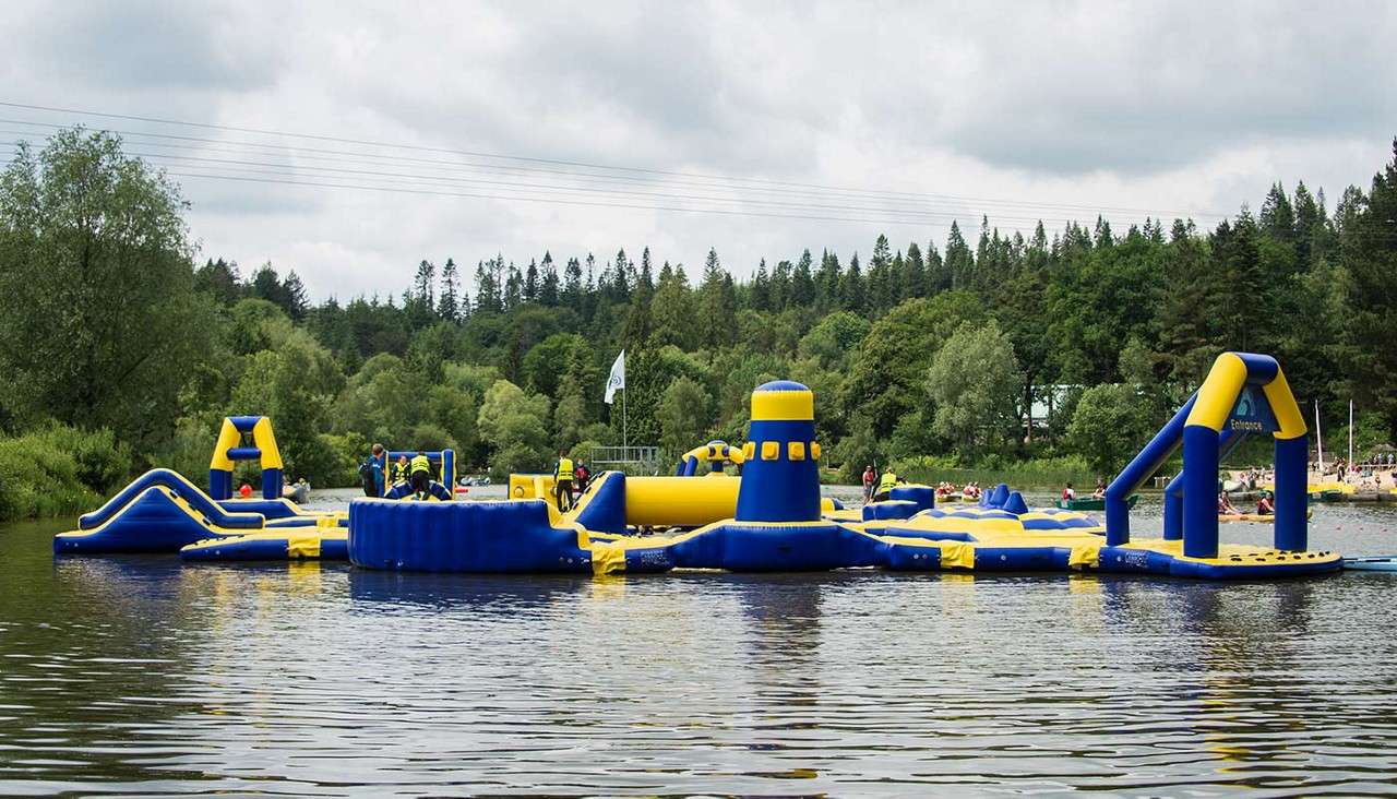 Inflatable water obstacle course hosts people in life jackets climbing and sliding, floating on a calm lake. Context: forested shoreline, scattered kayakers, overcast sky, and a beach in the distance.