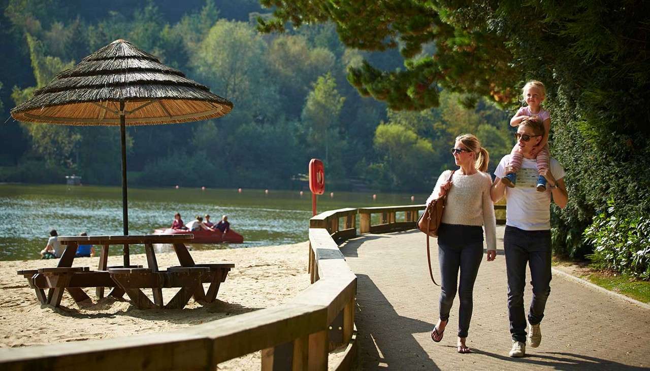 Family walks along a paved lakeside path; father carries a child on his shoulders while mother strolls beside, near a sandy beach with straw-parasol picnic table, pedal boaters on the lake, and surrounding forest.