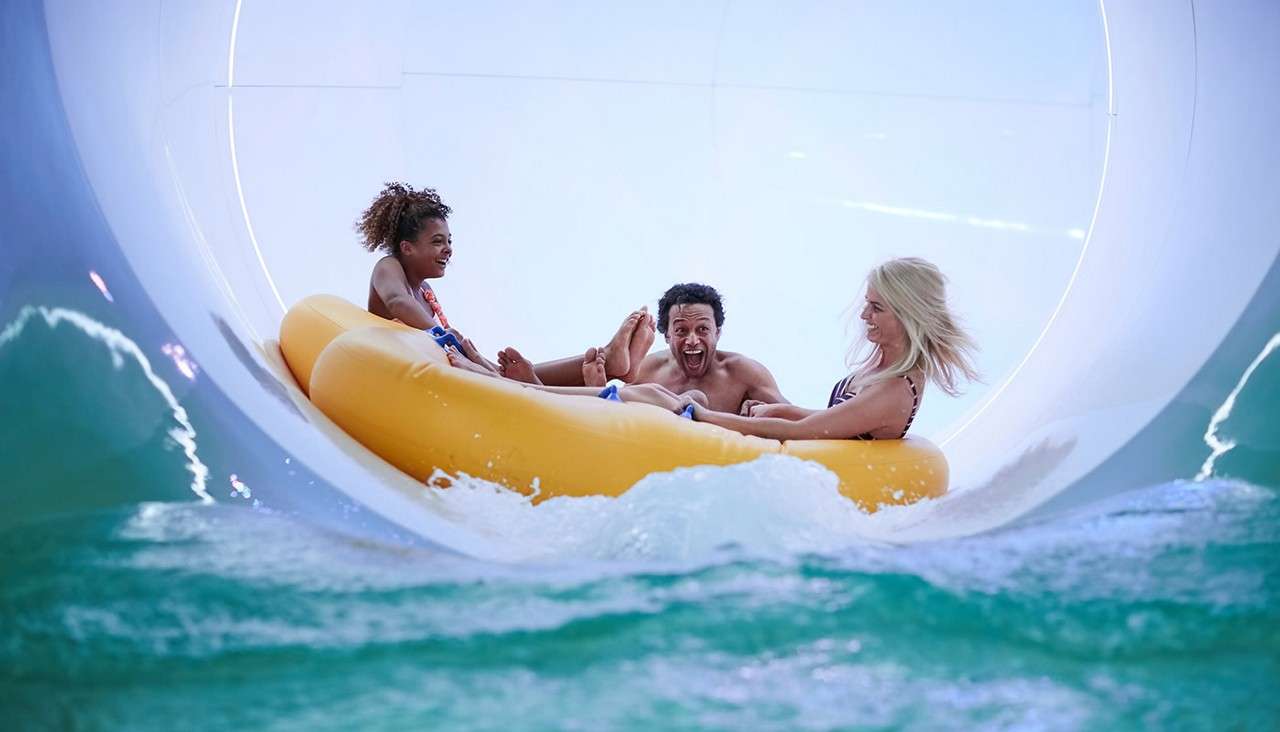 Three people on a yellow inflatable raft ride forward, laughing, inside a white enclosed water slide, as turquoise water splashes around the tube, suggesting a lively water park environment.