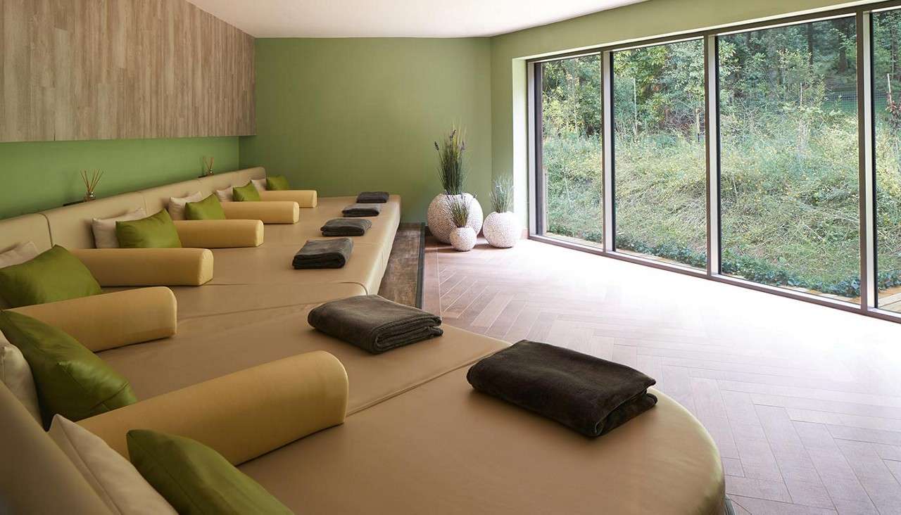 Beige lounge beds align, topped with gray towels and green/yellow bolsters; they invite rest. In a serene room with green walls, wood floor, potted plants, floor-to-ceiling windows overlooking forest.