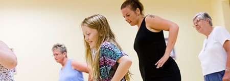 Group of people practice dance steps, hands on hips, leaning forward together, in a brightly lit studio with yellow walls; various ages line up, focusing on movement.