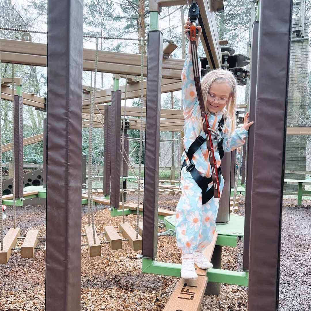 Child balances on a narrow wooden beam, gripping a safety lanyard while wearing a harness; surrounding ropes-course platforms and trees fill the background. Text on the beam: LIFT.