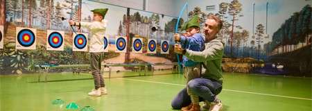 Two children in green hats practice archery with toy bows; an adult kneels, guiding one. Bullseye targets line the wall in an indoor range with forest mural and green floor.