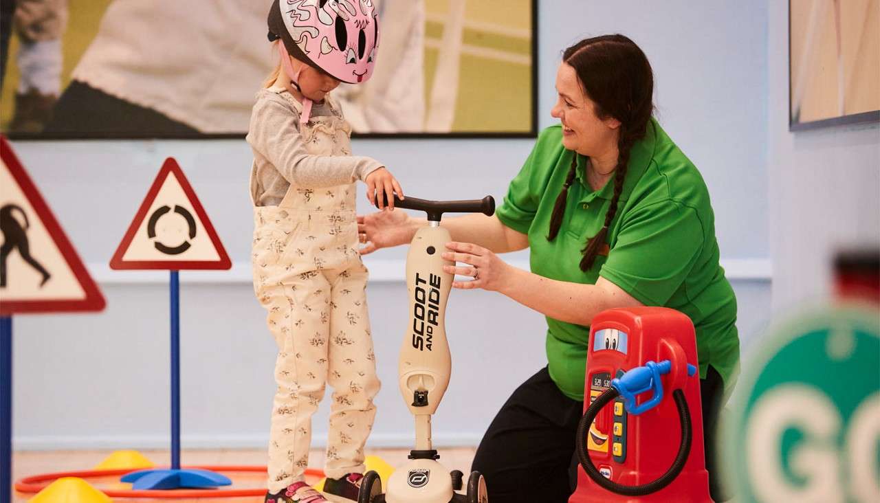 Child on a “SCOOT AND RIDE” scooter balances while an adult steadies them, in an indoor play area with toy traffic signs and a red gas pump; blurred green sign reads “GO.”