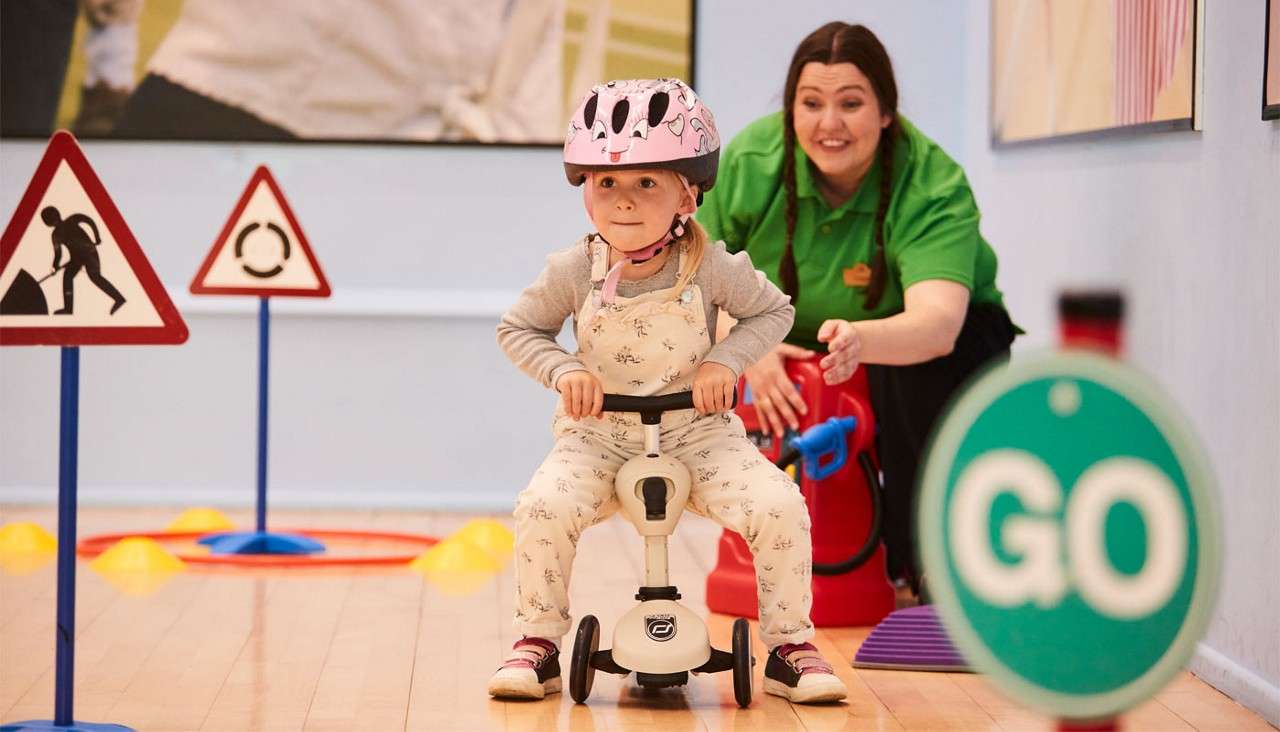 Child rides a small three-wheeled scooter, pedaling forward while an adult encourages behind; surrounded by indoor traffic training props—cones and signs, including a green circular sign reading: GO.