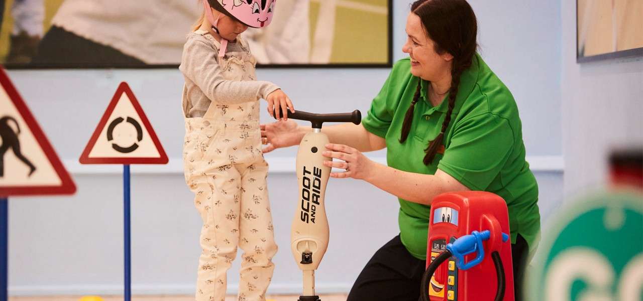 Child on a small scooter balances while an adult steadies them; indoor play area with traffic signs and a toy fuel pump. Text on scooter: SCOOT AND RIDE.