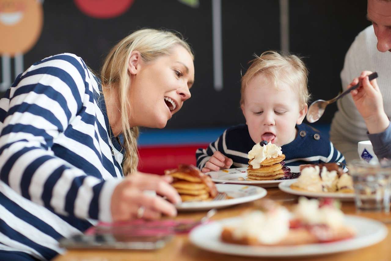 Child licks whipped cream–topped pancakes while adults eat and assist, seated at a restaurant table with multiple plates of pancakes and utensils; a casual, playful breakfast scene indoors.