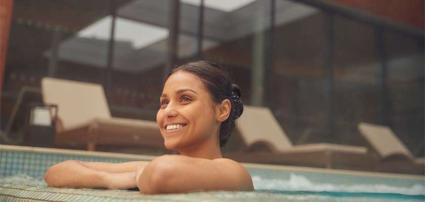 Person rests smiling with arms on pool edge, partially submerged, facing left. Indoor spa setting with tiled pool, blurred lounge chairs, and floor-to-ceiling windows under soft, diffused daylight.