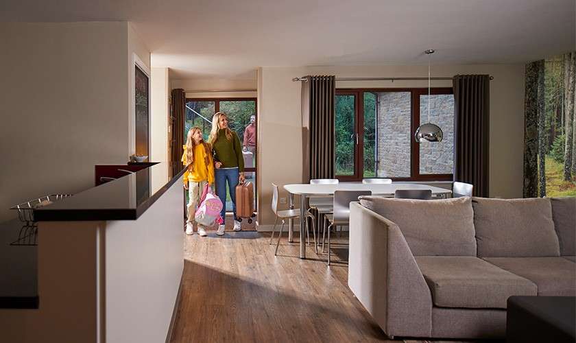 Family with suitcases enters a cabin; mother and child step in as another person holds the door. Open-plan living-dining room with gray sofa, wooden floor, large windows overlooking a forest.