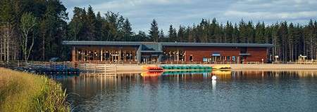 Lakeside boathouse sits beside a calm, reflective lake, displaying stacked colorful kayaks and paddleboats along a sandy shore, with docks and buoys, framed by dense conifer forest and evening light.