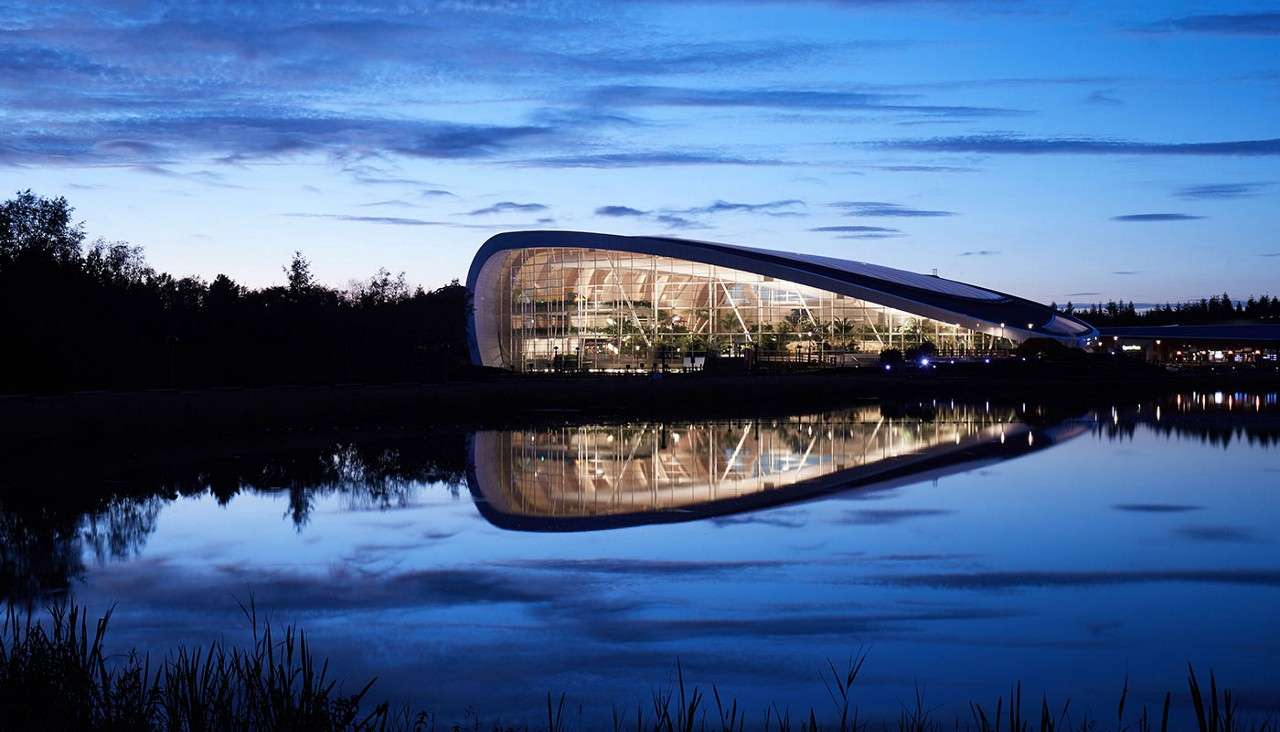 A sleek, illuminated modern building curves and glows, reflecting across a lake at dusk; framed by silhouetted trees, glass walls reveal interior structure and plants under blue twilight sky.
