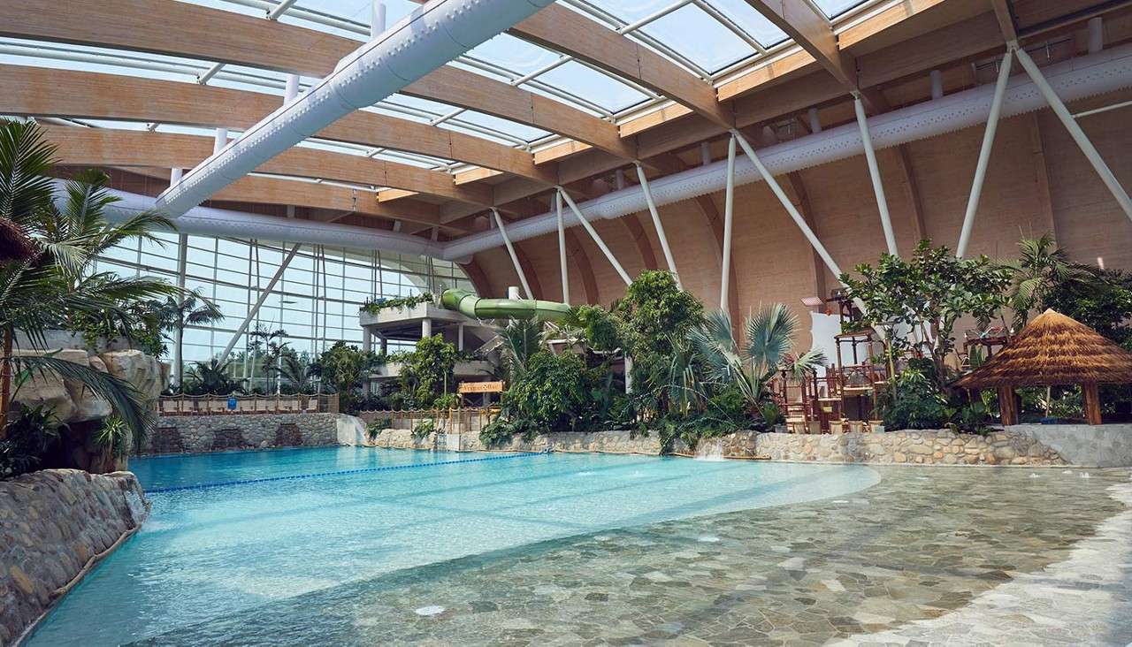 Indoor swimming pool glistens under skylights, bordered by rocks and tropical plants; nearby, a green waterslide and wooden play structures sit beneath a high arched roof with large glass windows.