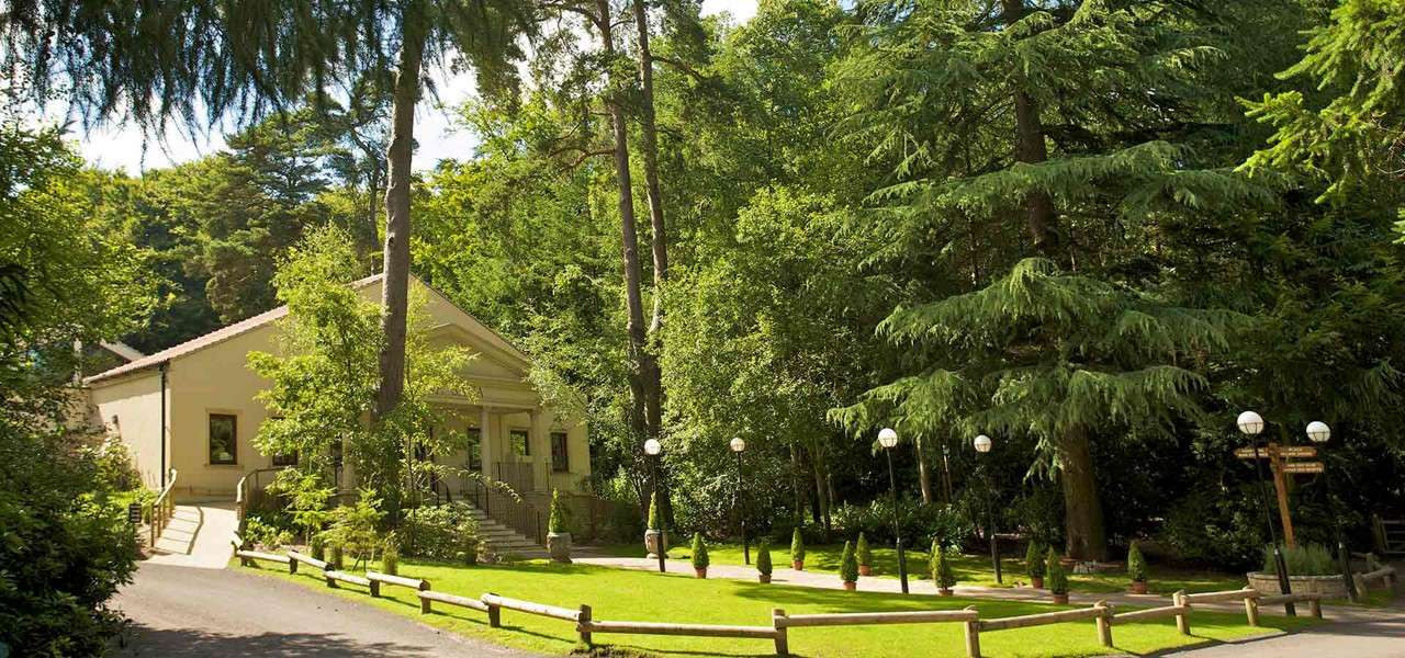 Cream-colored pavilion stands among conifers, front steps rising to a portico; round-globe lampposts and potted shrubs line a manicured lawn beside a rail and paved path in a wooded park.