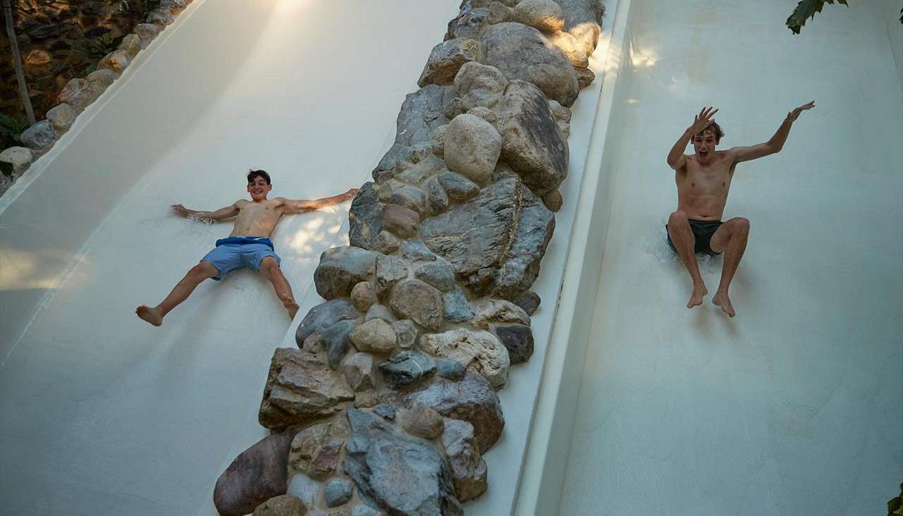 Two people slide down parallel white chutes, arms out, separated by a rocky divider, wearing swim trunks; they descend quickly in an outdoor setting with trees and dappled sunlight.