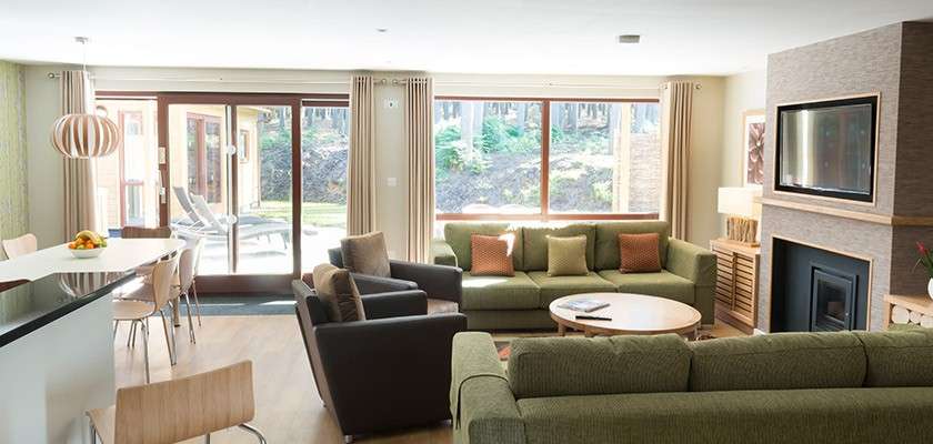 Sunlit living room features green sofas arranged around a round coffee table; sliding glass doors open to patio with loungers, while wall-mounted TV above fireplace faces kitchen island.