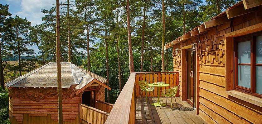 Wooden treehouse balcony holds a small lime-green table and chairs, overlooking another cabin amid tall pine trees, with sunlight filtering through a forest setting.