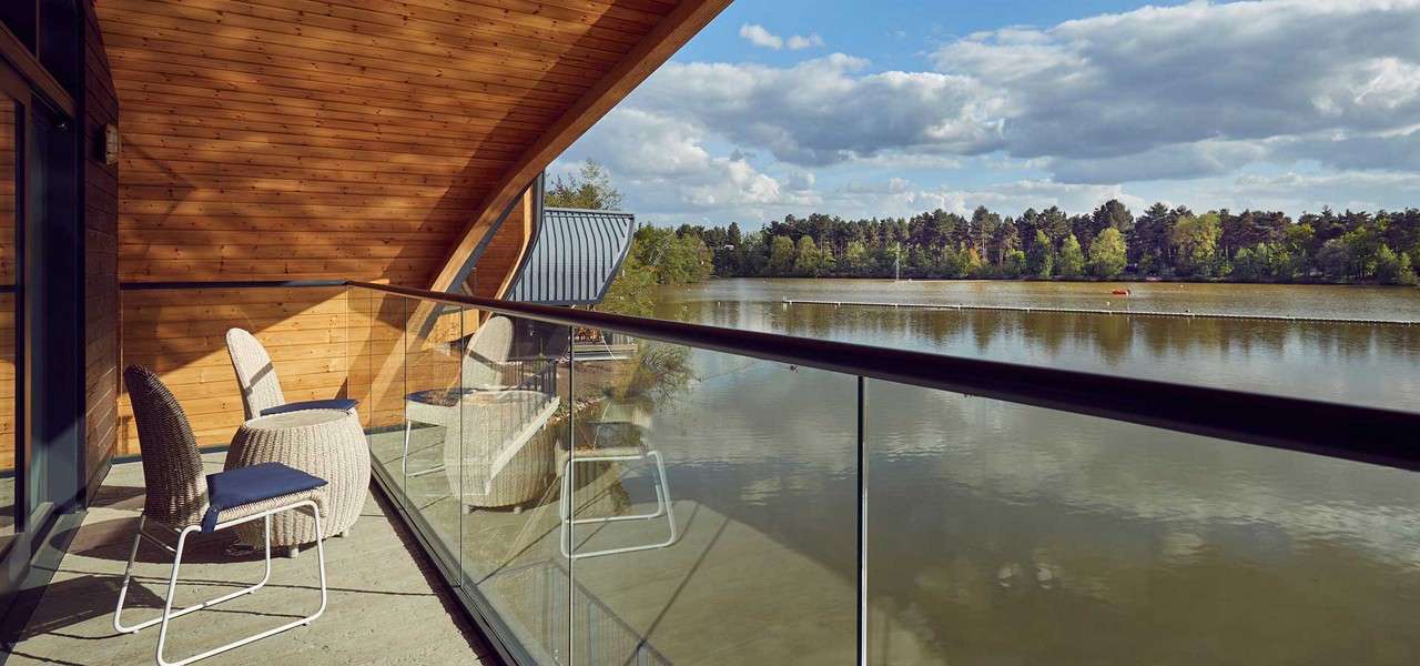 Balcony chairs sit empty, facing a calm lake behind a glass railing. Curved wooden walls and ceiling shelter the space; across the water, a tree-lined shore rests under scattered clouds.