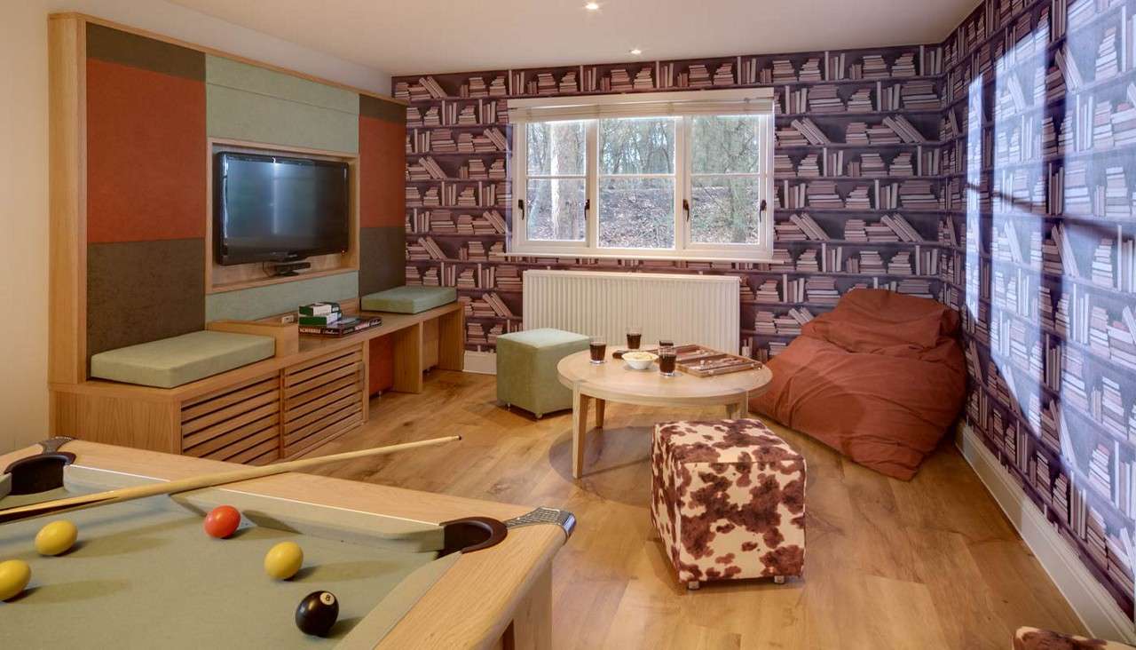 Pool table holds balls and cues, foregrounding a furnished recreation room with wall-mounted TV, round coffee table with drinks, ottomans, beanbag chair, wood floor, window, and bookshelf-patterned wallpaper.