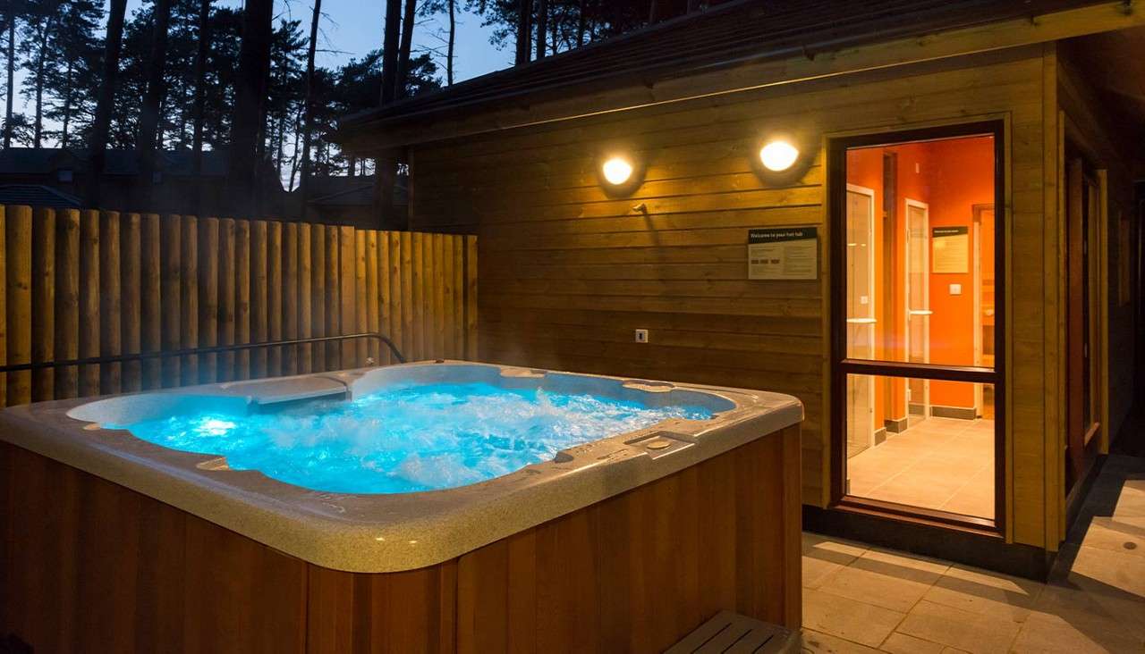 Hot tub bubbling and steaming with blue light beside a wood cabin. Wall lamps glow; open doorway shows interior. Wooden fence and tall trees at dusk. Signs with unreadable text.