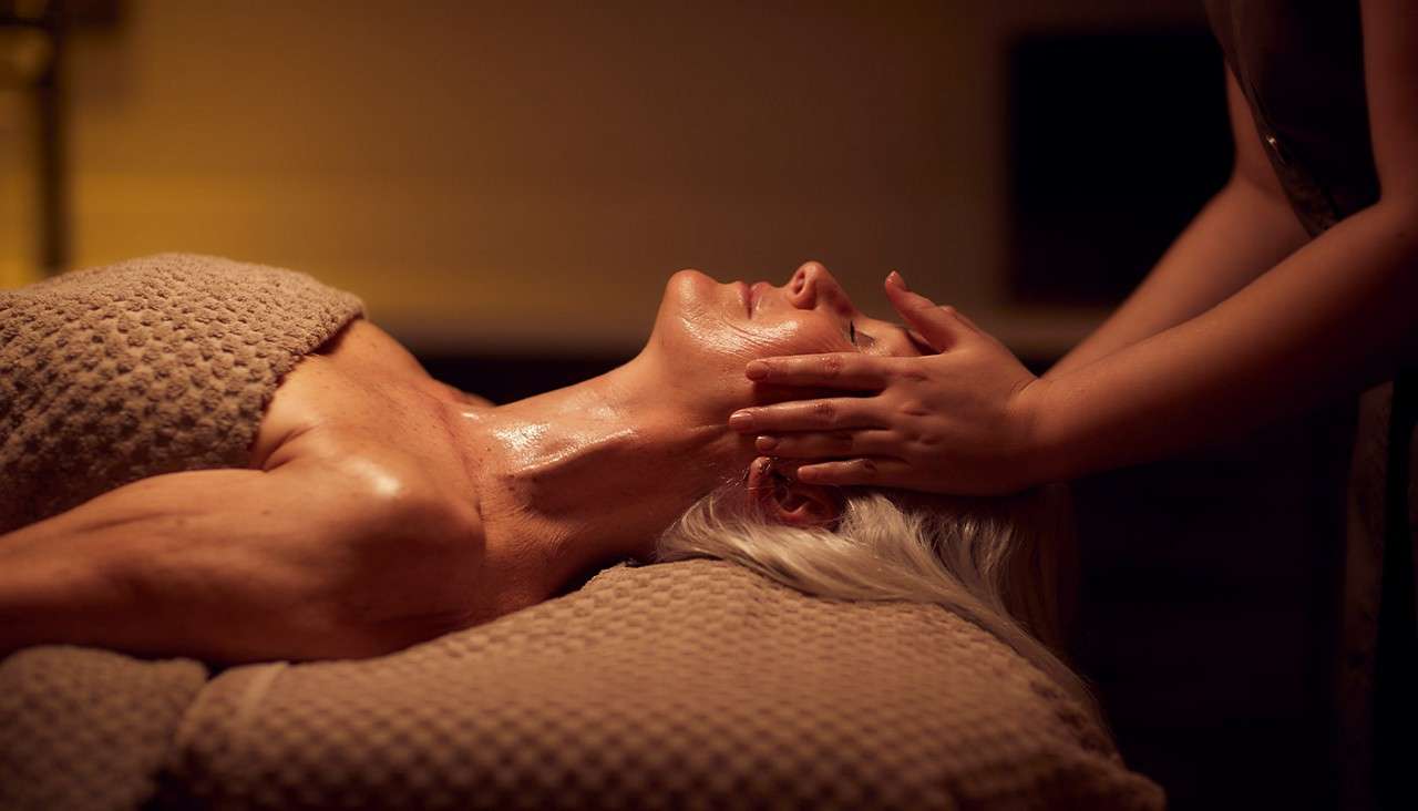 Person receiving a face and neck massage lies on a padded table; hands gently press temples; oiled skin glistens in warm, dim spa lighting.