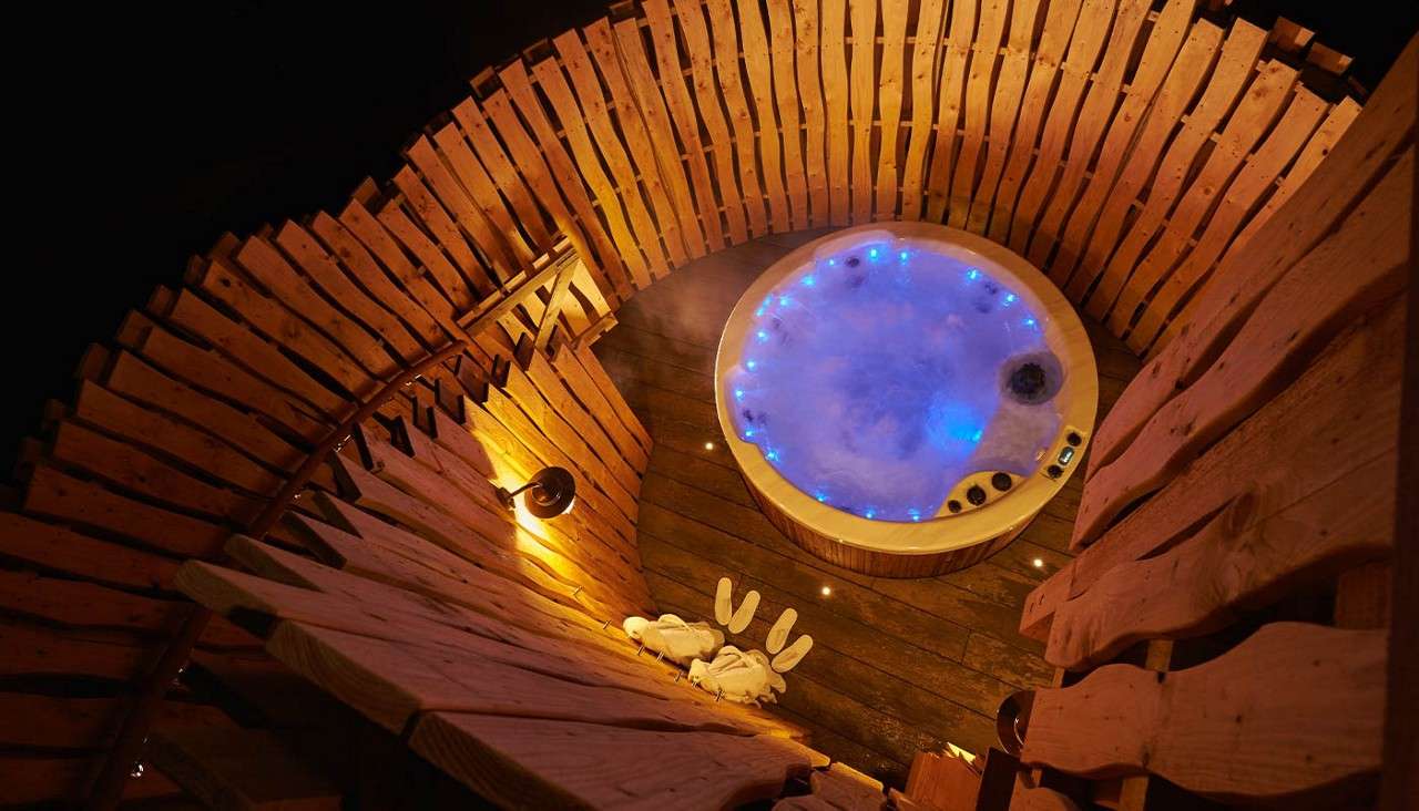 Bubbling circular hot tub glows blue, steaming. Surrounded by a rustic wooden enclosure and deck, a warm wall lamp shines; white robes and slippers rest nearby, at night.