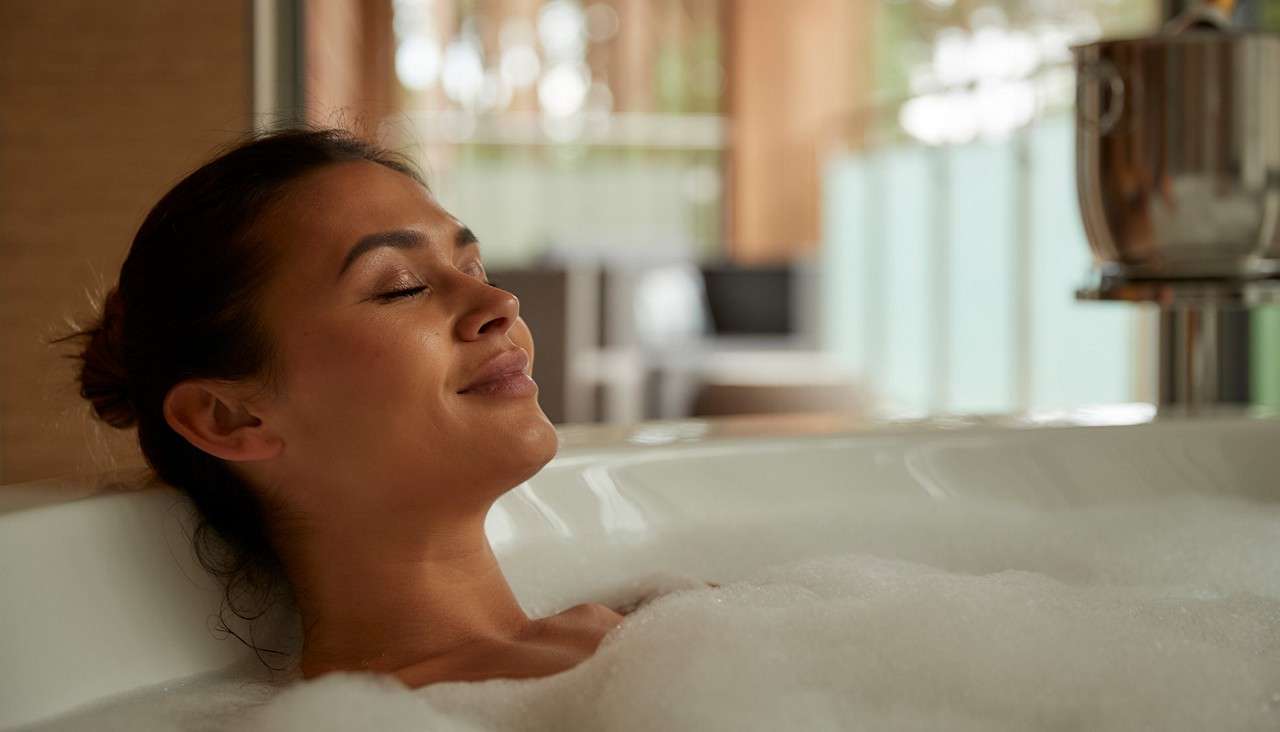 A person reclines in a bubble bath, eyes closed and smiling, surrounded by foam; a modern, softly lit bathroom with blurred furnishings and a metal ice bucket in the background.