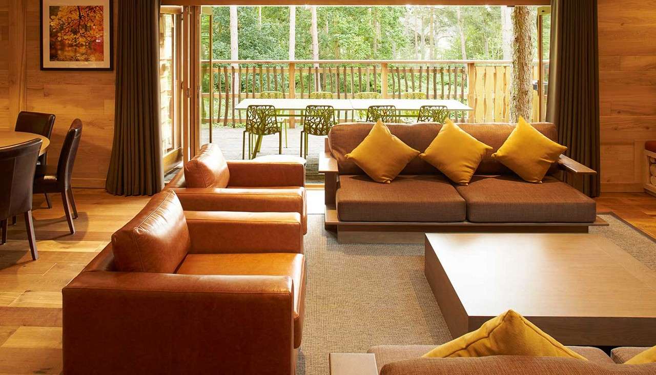 Brown leather armchairs and a sofa sit around a low wooden coffee table, facing open sliding doors; beyond, a deck with green chairs overlooks a wooded forest.