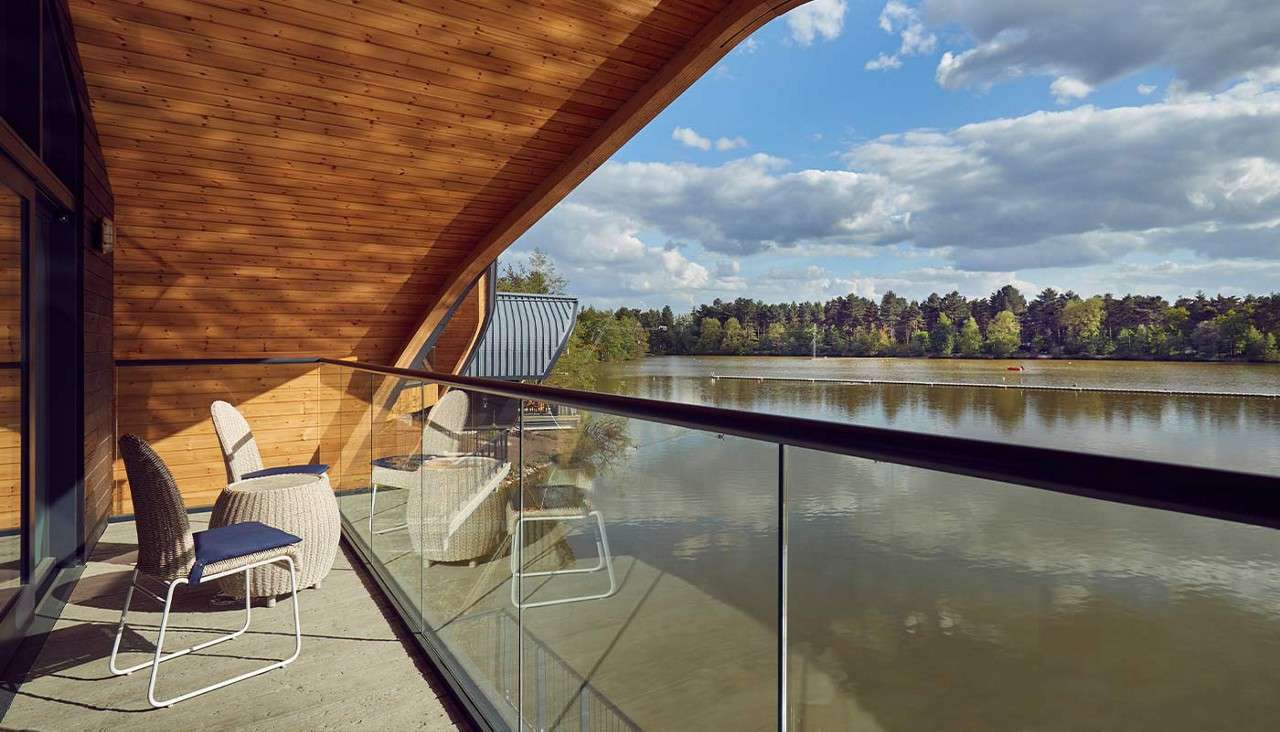 Balcony seating overlooks a calm lake. Two wicker chairs and a small table sit under a curved timber canopy behind a glass railing, facing water, tree-lined shore, and cloudy sky.