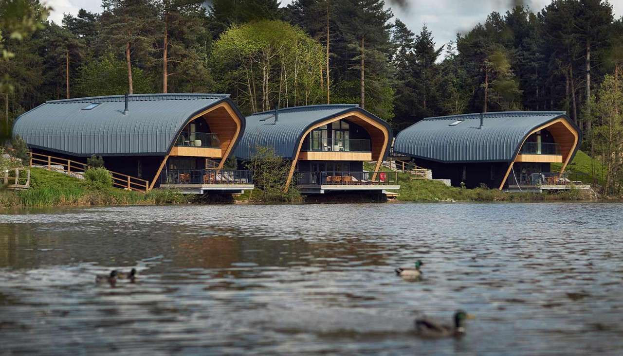 Three modern lakeside lodges extend over the water on stilts, featuring curved metal roofs and glass balconies; ducks swim in the foreground, while a dense pine forest and cloudy sky surround.