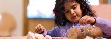 Child arranges small items on a log, concentrating at a table; cotton-like clumps and pebbles surround her in a softly lit indoor classroom setting.