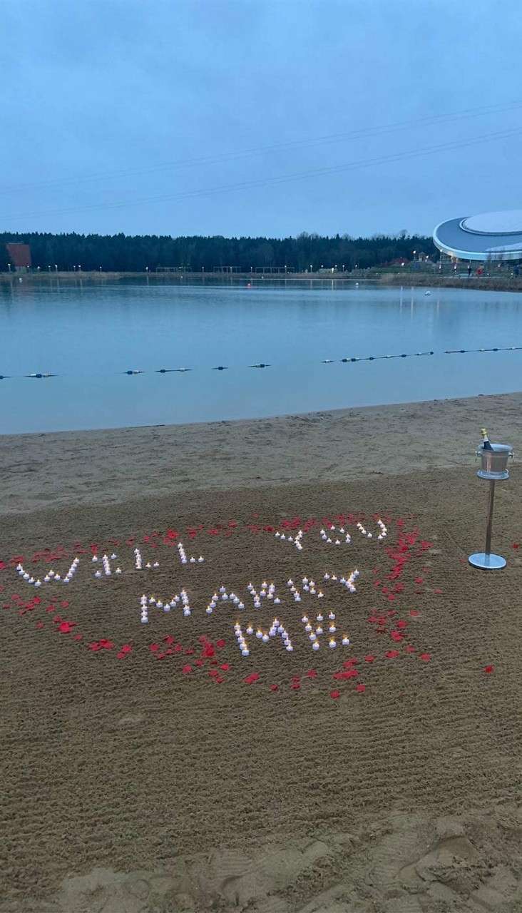 Candles spell "WILL YOU MARRY ME" on a beach, ringed by red petals; a champagne bottle in an ice bucket on a stand; a lakeside setting at dusk.