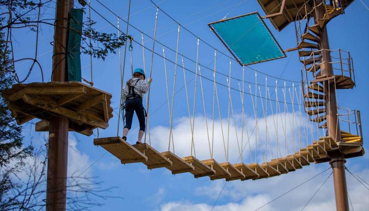 A harnessed climber crosses a suspended wooden plank bridge, holding safety ropes, between elevated platforms; spiral stairs and nets nearby, against a blue sky with clouds and trees.