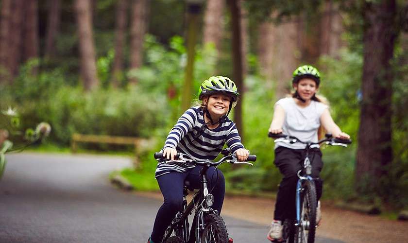 Two children ride bicycles, smiling and wearing helmets, along a winding paved path; trees and greenery surround them in a forested park.