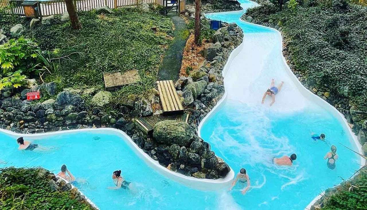 Swimmers drift and slide along a winding, shallow blue-water channel, bordered by rocks and dense greenery in an outdoor park. A small red sign bears illegible white text.