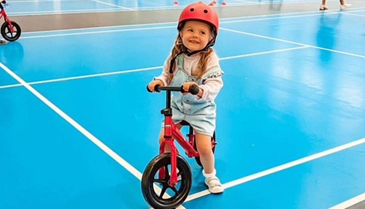 Child on a red balance bike smiles while gliding, wearing a red helmet and denim overalls; indoors on a bright blue sports court with white lines and distant cones.