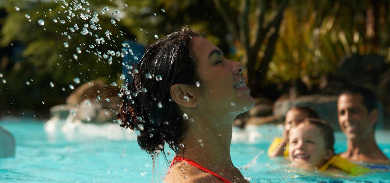 Person in red swimsuit tilts head back, water droplets splashing upward; in background, adults and child with yellow floaties wade in a tropical-style swimming pool surrounded by greenery.