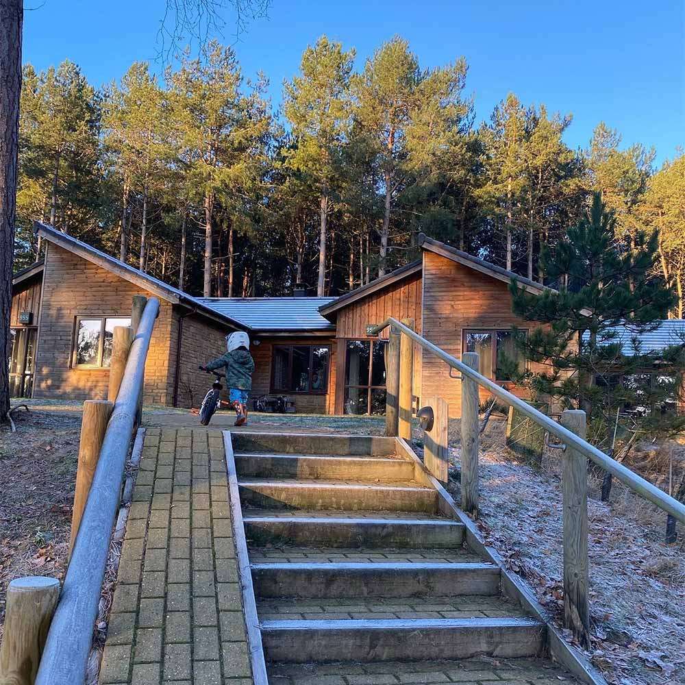 Child wearing helmet pushes a balance bike up a ramp beside steps, approaching wooden cabins. Pine forest surrounds the cabins; frosty ground and blue sky suggest a cold morning.