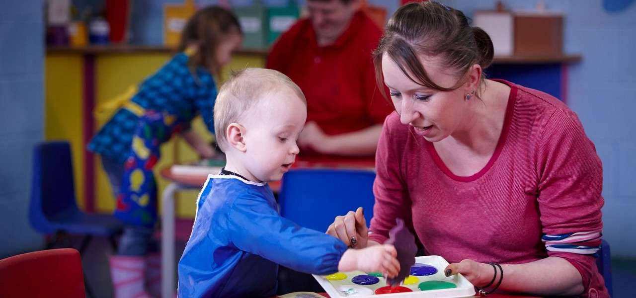 Toddler, guided by an adult, dips a sponge into colorful paint on a palette and stamps paper; other children and an adult work nearby in a bright classroom.