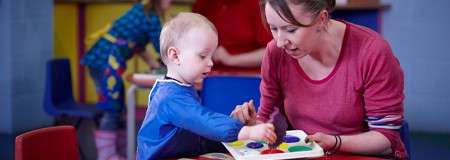 Child finger-paints with an adult, dabbing colors on a palette; they sit at a small table. Background shows a colorful classroom with shelves, chairs, and another child playing.