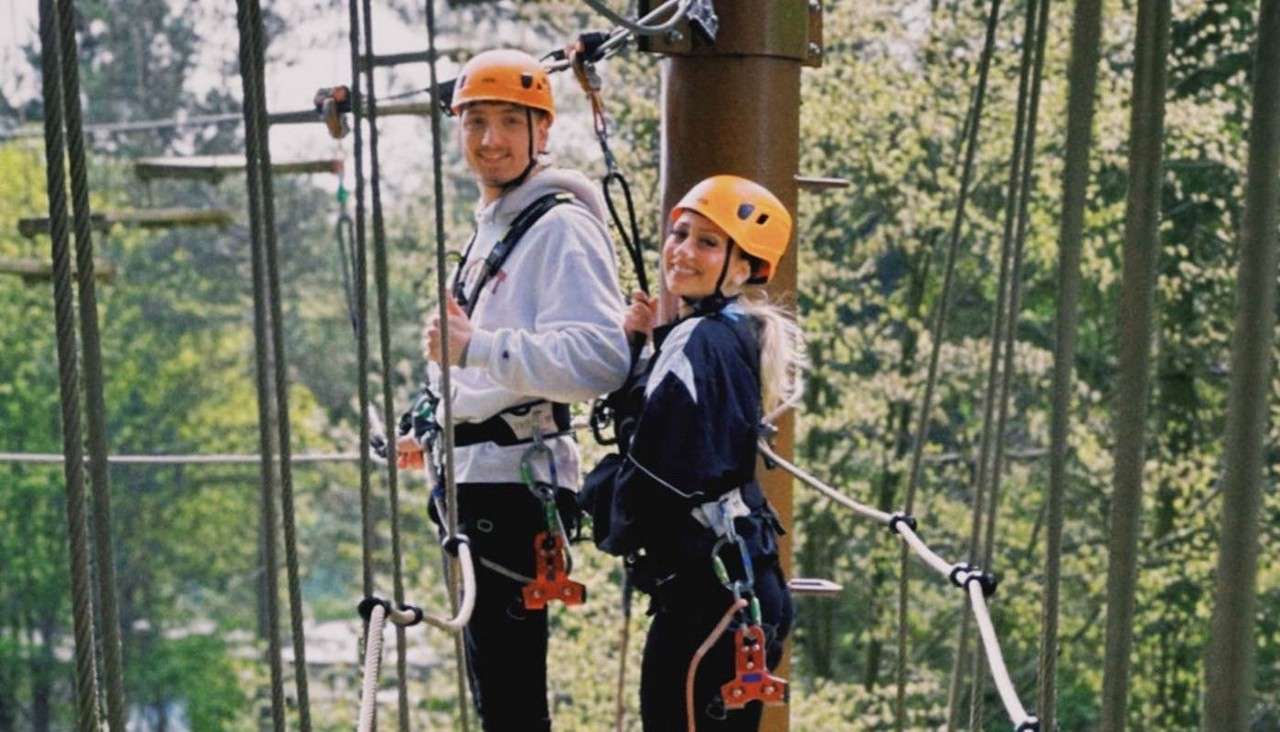 Two harnessed people clip onto safety lines, crossing a ropes-course platform; they wear orange helmets and jackets, smiling, surrounded by tall trees in a forest canopy.