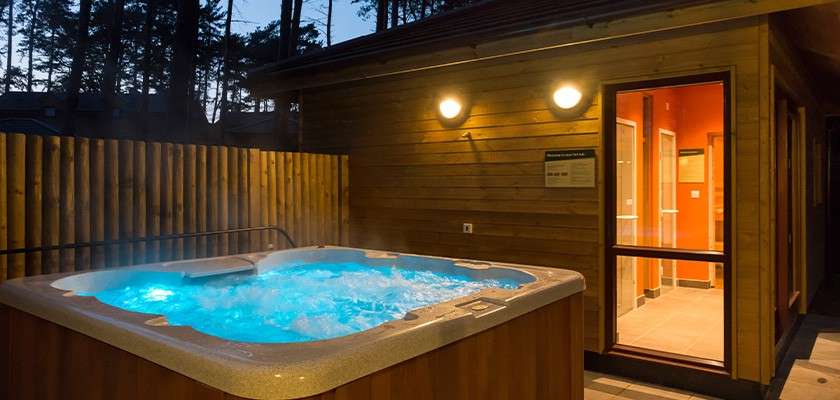 Hot tub bubbles and steams, glowing blue. It sits on a wooden patio beside a lit cabin entrance, surrounded by a tall timber fence and silhouetted pine trees at dusk.