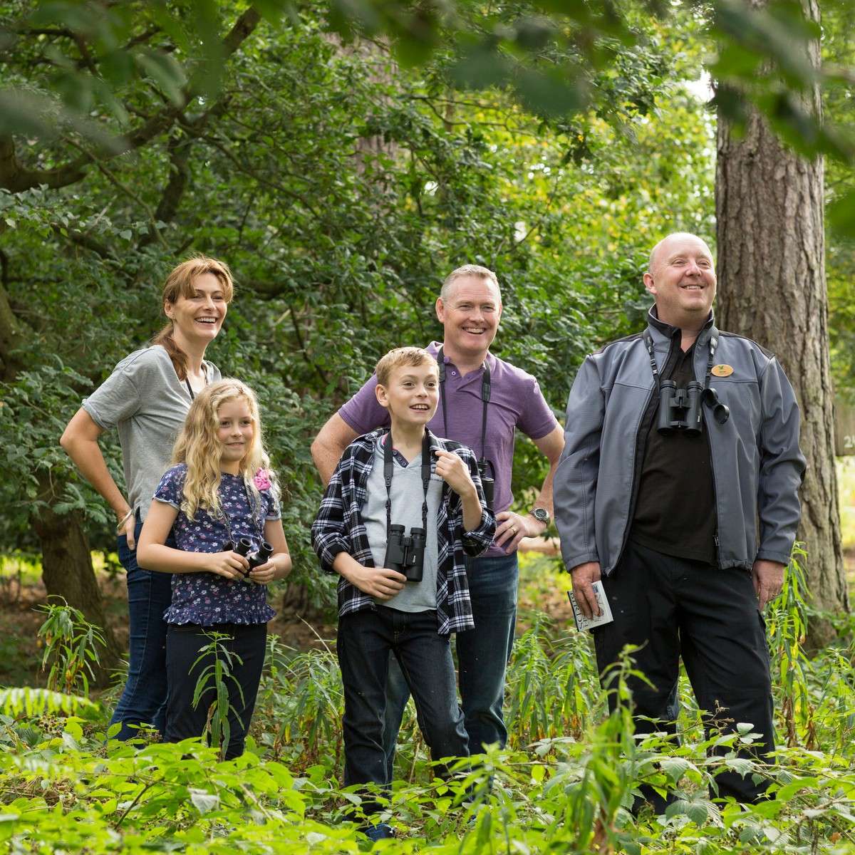 Group of five birdwatchers stand together, smiling and looking upward, holding binoculars; context: lush green forest with trees and undergrowth during a daylight hike.
