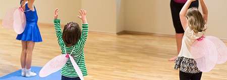 Children wearing pink fairy wings raise their arms while following an instructor, standing on a wooden floor in a dance studio; one child on a blue mat, others spaced apart.